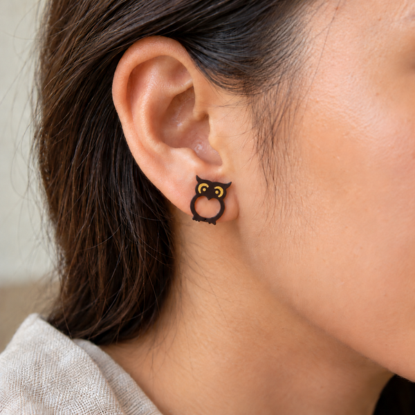 Dark brown owl stud earrings with yellow eye details, an inward-pointed opening and asymmetric feet, worn by an adult woman